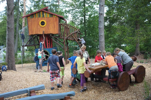 buntes Baumhaus aus Holz mit Kletter- und Spielmöglichkeiten, davor ein Tisch mit Bänken; Kinder und Erwachsene beim Picknick und Spiel